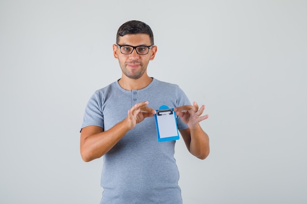 Foto gratuita hombre joven en camiseta gris, gafas sosteniendo mini portapapeles y mirando contento