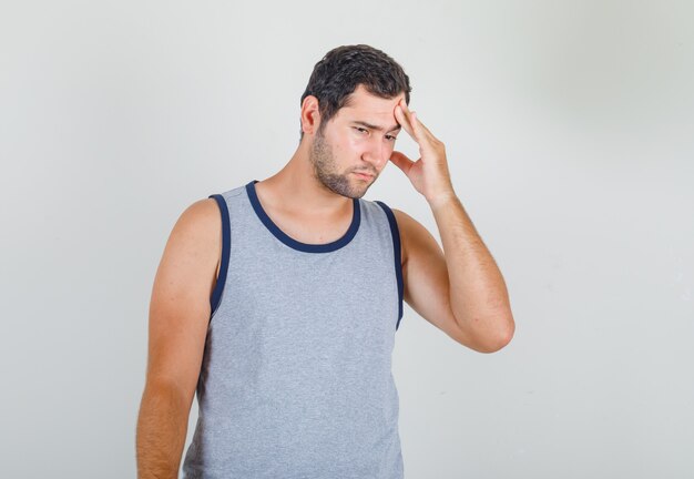 Hombre joven en camiseta gris que muestra cuánto le duele la cabeza y parece miserable