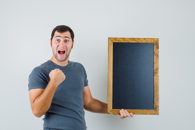 Foto gratuita hombre joven en camiseta gris sosteniendo pizarra y mirando feliz