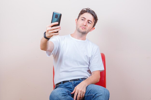 Hombre joven en camiseta, jeans tomando selfie en teléfono móvil mientras está sentado en una silla