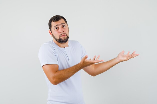 Hombre joven en camiseta mostrando gesto de impotencia y mirando confundido, vista frontal.