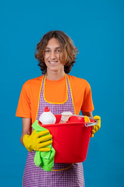 Hombre joven en camiseta naranja con delantal guantes de goma sosteniendo un cubo con herramientas de limpieza mirando a la cámara sonriendo positivo y feliz de pie sobre fondo azul.