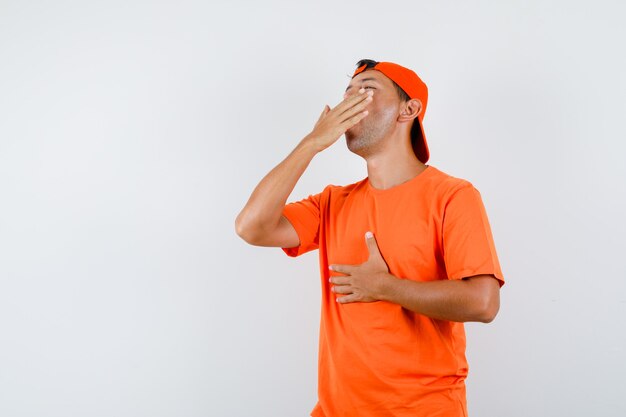 Hombre joven en camiseta naranja y gorra bostezando y mirando soñoliento