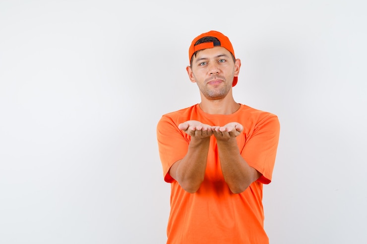 Foto gratuito hombre joven en camiseta naranja y gorra manteniendo las manos juntas como presentando algo y luciendo suave
