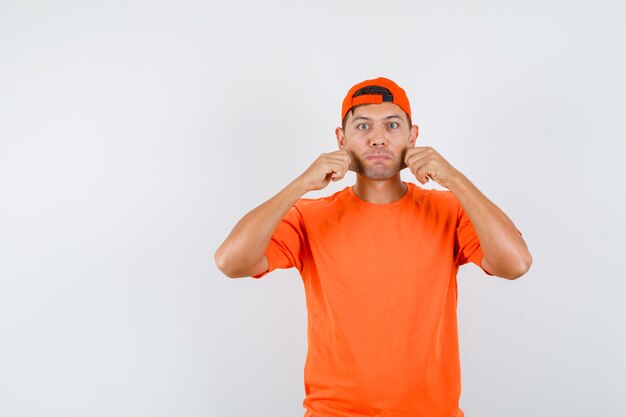 Hombre joven en camiseta naranja y gorra tirando de sus mejillas y mirando amado