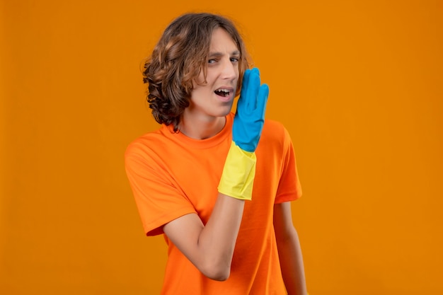 Foto gratuita hombre joven en camiseta naranja con guantes de goma contando un secreto con la mano cerca de la boca mirando confiado de pie sobre fondo amarillo