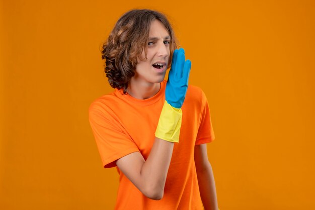Hombre joven en camiseta naranja con guantes de goma contando un secreto con la mano cerca de la boca mirando confiado de pie sobre fondo amarillo