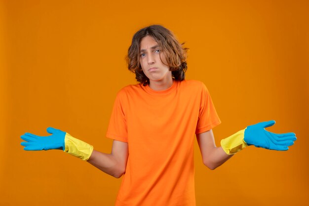 Hombre joven en camiseta naranja con guantes de goma encogiéndose de hombros mirando incierto y confundido sin respuesta extendiendo las palmas de pie sobre fondo amarillo