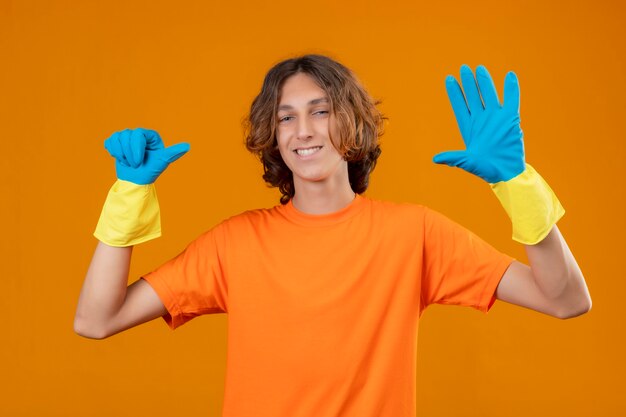 Hombre joven en camiseta naranja con guantes de goma sonriendo con cara feliz mostrando y apuntando hacia arriba con los dedos número seis de pie sobre fondo amarillo