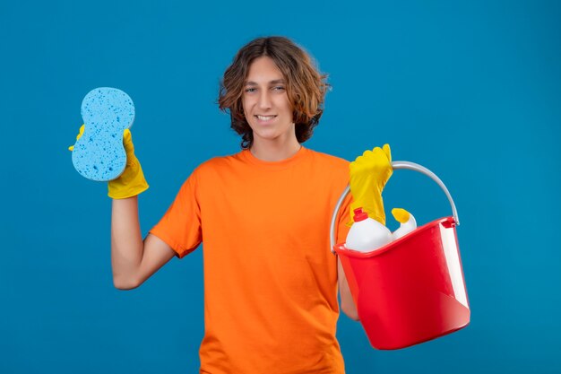 Hombre joven en camiseta naranja con guantes de goma sosteniendo un balde con herramientas de limpieza y una esponja mirando a la cámara sonriendo alegremente feliz y positivo de pie sobre fondo azul