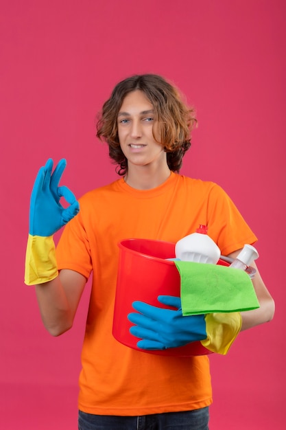 Hombre joven en camiseta naranja con guantes de goma sosteniendo un balde con herramientas de limpieza mirando a la cámara sonriendo confiado haciendo bien firmar sobre fondo rosa