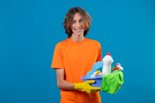 Hombre joven en camiseta naranja con guantes de goma sosteniendo un balde con herramientas de limpieza mirando a la cámara sonriendo positivo y feliz de pie sobre fondo azul