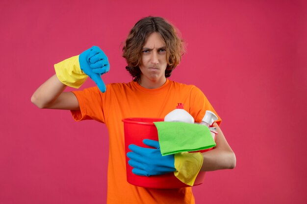 Hombre joven en camiseta naranja con guantes de goma sosteniendo un balde con herramientas de limpieza mirando disgustado mostrando los pulgares hacia abajo con el ceño fruncido sobre fondo rosa