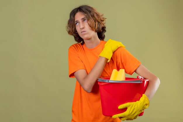 Hombre joven en camiseta naranja con guantes de goma sosteniendo un balde con herramientas de limpieza mirando mal tocar el hombro con dolor de pie sobre fondo verde