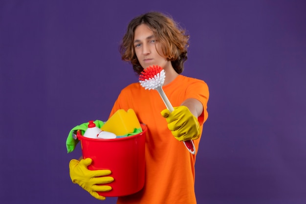 Hombre joven en camiseta naranja con guantes de goma sosteniendo un balde con herramientas de limpieza que muestra un cepillo para fregar a la cámara mirando confiado de pie sobre fondo púrpura