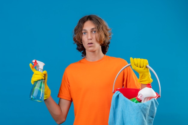 Hombre joven en camiseta naranja con guantes de goma sosteniendo un balde con herramientas de limpieza y spray de limpieza mirando a la cámara sorprendido de pie sobre fondo azul.