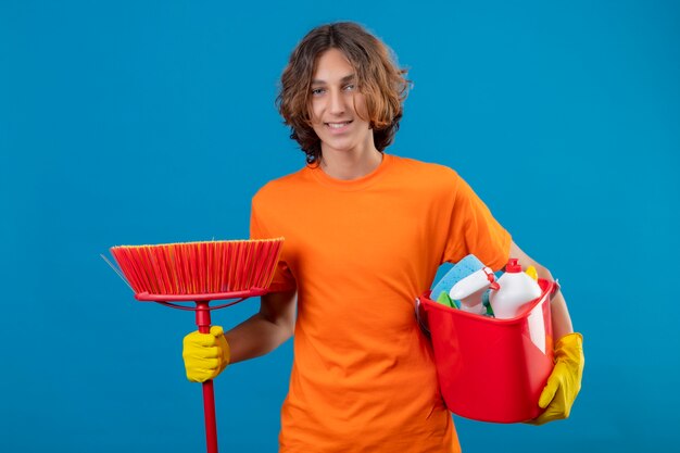 Foto gratuita hombre joven en camiseta naranja con guantes de goma sosteniendo un balde con herramientas de limpieza y un trapeador mirando a la cámara sonriendo positivo y feliz de pie sobre fondo azul.