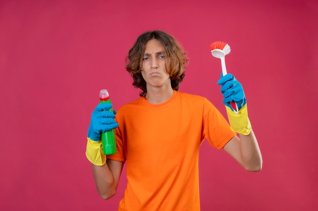 Hombre joven en camiseta naranja con guantes de goma sosteniendo una botella de productos de limpieza y un cepillo para fregar mirando a la cámara disgustado con el ceño fruncido sobre fondo rosa