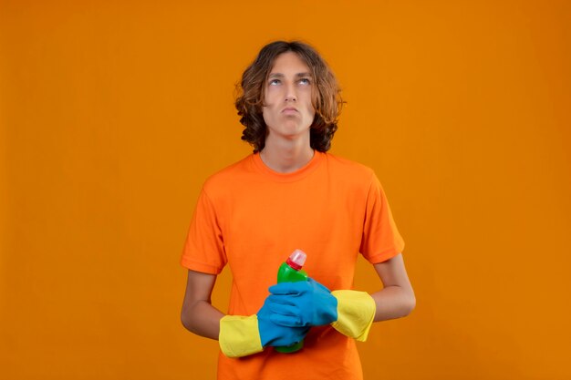 Hombre joven en camiseta naranja con guantes de goma sosteniendo una botella de productos de limpieza mirando hacia arriba con expresión pensativa de pie sobre fondo amarillo