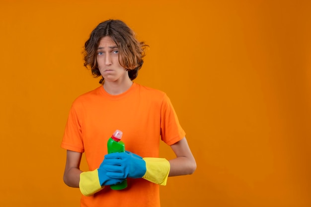 Foto gratuita hombre joven en camiseta naranja con guantes de goma sosteniendo una botella de productos de limpieza mirando confundido y muy ansioso de pie sobre fondo amarillo