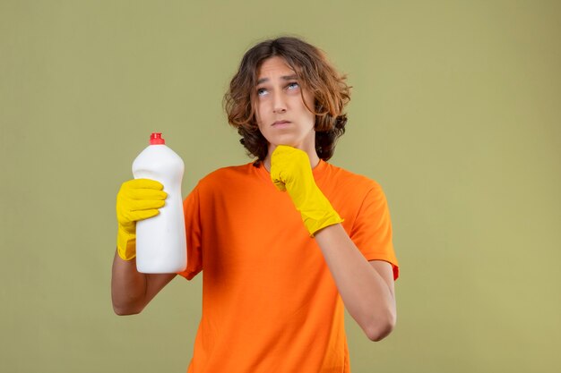 Hombre joven en camiseta naranja con guantes de goma sosteniendo una botella de productos de limpieza de pie con la mano en la barbilla mirando hacia arriba con expresión pensativa pensando sobre fondo verde