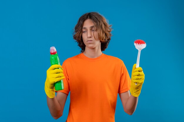 Hombre joven en camiseta naranja con guantes de goma sosteniendo un cepillo y una botella con productos de limpieza que parecen inciertos teniendo dudas sobre fondo azul