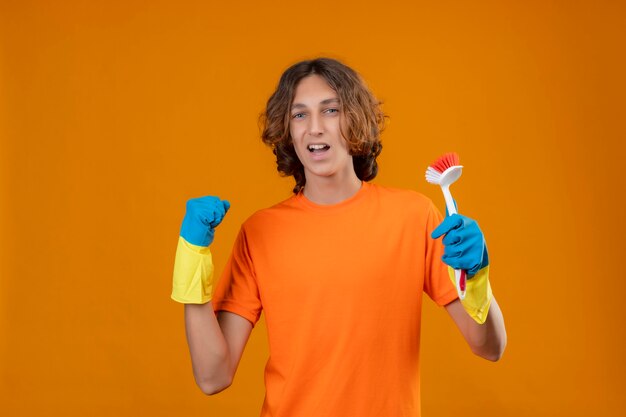 Hombre joven en camiseta naranja con guantes de goma sosteniendo un cepillo de fregar apretando el puño regocijándose de su éxito y victoria salido y feliz de pie sobre fondo amarillo