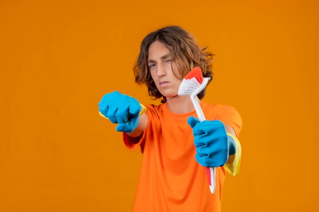 Hombre joven en camiseta naranja con guantes de goma sosteniendo cepillo de fregar apuntando a la cámara con expresión sospechosa en la cara de pie sobre fondo amarillo