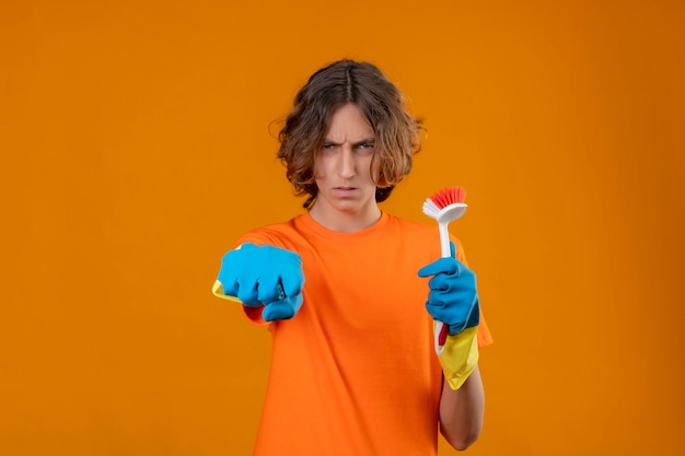 Hombre joven en camiseta naranja con guantes de goma sosteniendo cepillo de fregar apuntando a la cámara con expresión sospechosa en la cara de pie sobre fondo amarillo