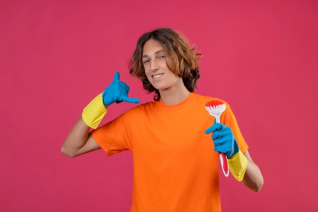 Hombre joven en camiseta naranja con guantes de goma sosteniendo un cepillo de fregar mirando a la cámara con una sonrisa de confianza haciendo gesto de llamarme de pie sobre fondo rosa