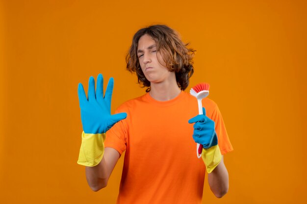 Hombre joven en camiseta naranja con guantes de goma sosteniendo cepillo de fregar de pie con la mano abierta haciendo gesto de defensa de señal de stop sobre fondo amarillo