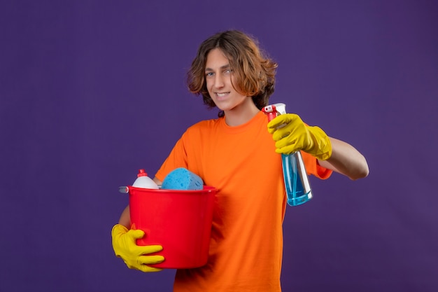 Hombre joven en camiseta naranja con guantes de goma sosteniendo un cubo con herramientas de limpieza y spray de limpieza mirando a la cámara con una sonrisa de confianza en la cara de pie sobre fondo púrpura
