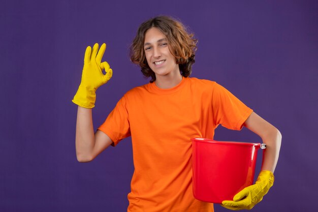 Hombre joven en camiseta naranja con guantes de goma sosteniendo un cubo sonriendo alegremente mirando a la cámara feliz y positivo haciendo bien signo de pie sobre fondo púrpura