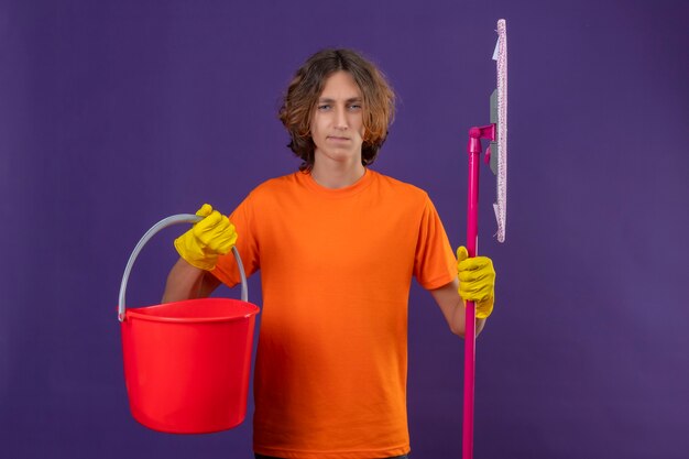 Hombre joven en camiseta naranja con guantes de goma sosteniendo un cubo y un trapeador mirando a la cámara con expresión seria y segura de pie sobre fondo púrpura