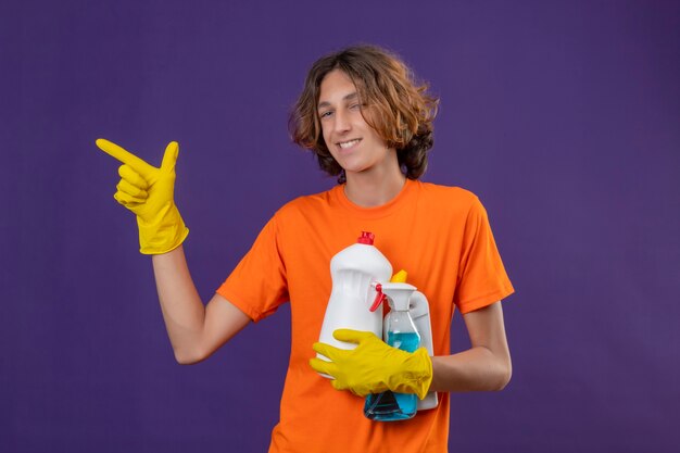 Hombre joven en camiseta naranja con guantes de goma sosteniendo herramientas de limpieza apuntando hacia el lado sonriendo alegremente de pie sobre fondo púrpura