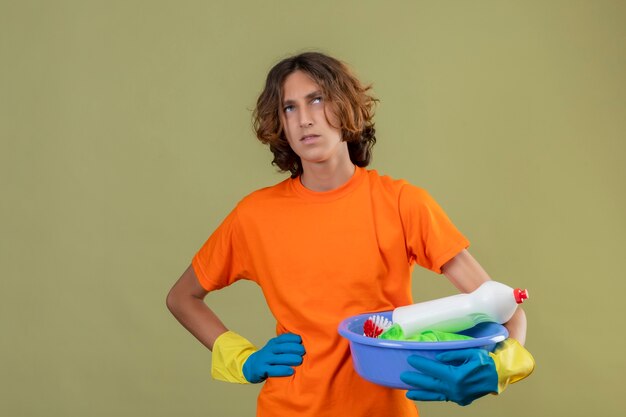 Hombre joven en camiseta naranja con guantes de goma sosteniendo el lavabo con herramientas de limpieza mirando hacia arriba con expresión pensativa en la cara pensando de pie sobre fondo verde