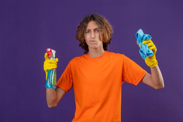 Hombre joven en camiseta naranja con guantes de goma sosteniendo spray de limpieza y alfombra mirando a la cámara con expresión escéptica en la cara de pie sobre fondo púrpura