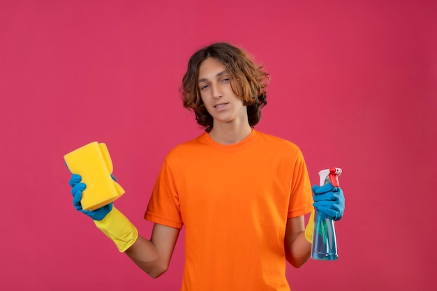 Hombre joven en camiseta naranja con guantes de goma sosteniendo spray de limpieza y una esponja mirando a la cámara con una sonrisa segura de pie sobre fondo rosa