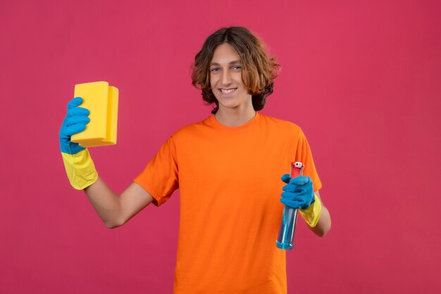 Hombre joven en camiseta naranja con guantes de goma sosteniendo spray de limpieza y una esponja mirando a la cámara con una sonrisa segura de pie sobre fondo rosa