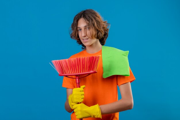 Hombre joven en camiseta naranja con guantes de goma sosteniendo un trapeador y una alfombra mirando a la cámara con una sonrisa de confianza en la cara de pie sobre fondo azul.