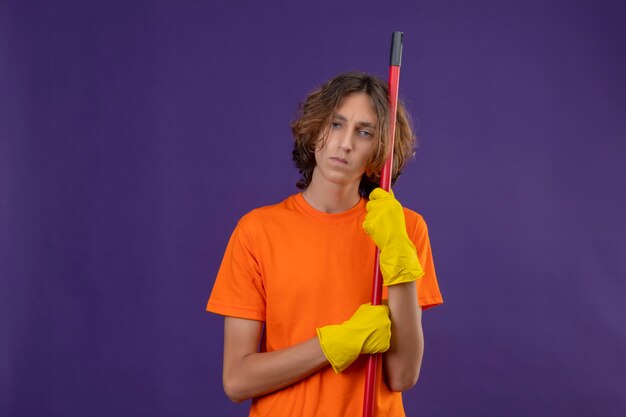 Hombre joven en camiseta naranja con guantes de goma sosteniendo un trapeador mirando a la cámara con expresión triste en la cara de pie sobre fondo púrpura