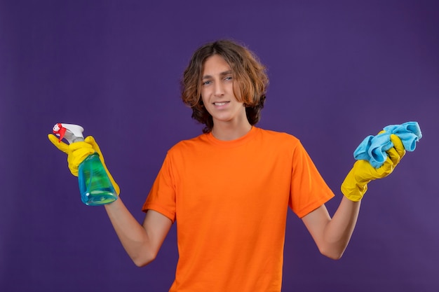 Hombre joven en camiseta naranja con guantes de goma con spray de limpieza y alfombra mirando a la cámara con una sonrisa en la cara listo para limpiar de pie sobre fondo púrpura