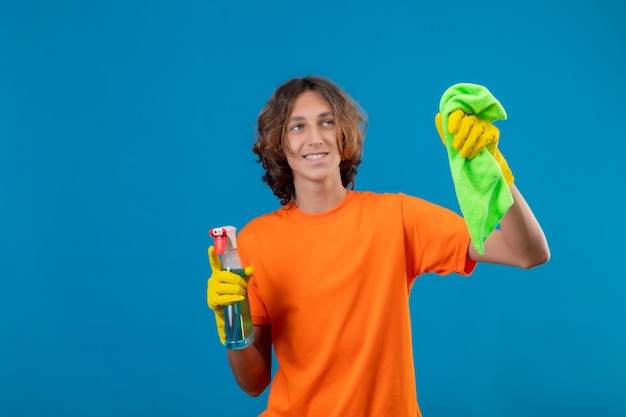 Foto gratuita hombre joven en camiseta naranja con guantes de goma con spray de limpieza y alfombra mirando a la cámara con una sonrisa de confianza listo para limpiar de pie sobre fondo azul.