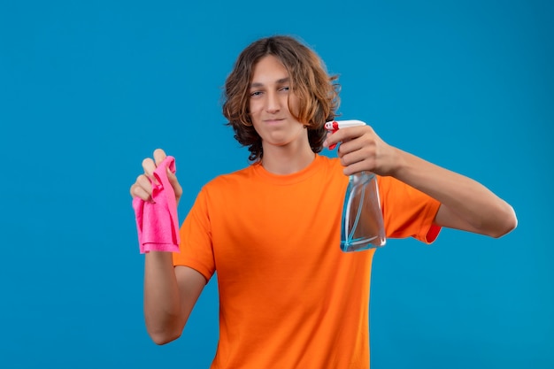 Hombre joven en camiseta naranja con guantes de goma con spray de limpieza y alfombra mirando a la cámara con una sonrisa de confianza listo para limpiar de pie sobre fondo azul.