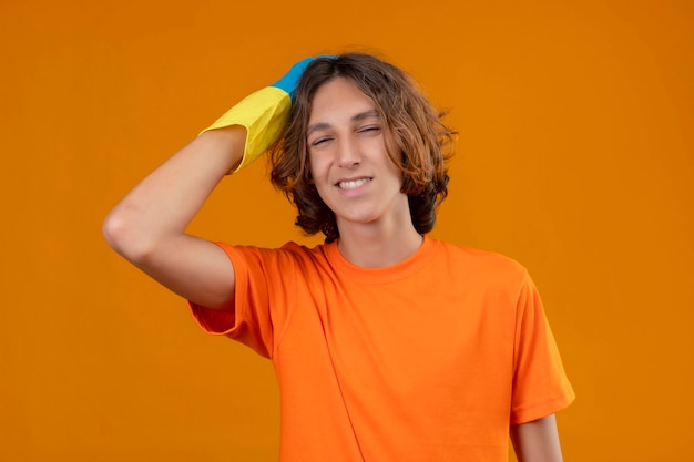 Hombre joven en camiseta naranja con guantes de goma tocando la cabeza por error mirando confundido concepto de mala memoria sobre fondo amarillo