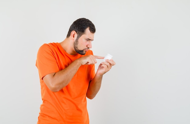 Hombre joven en camiseta naranja leyendo información sobre botella de píldoras, vista frontal.