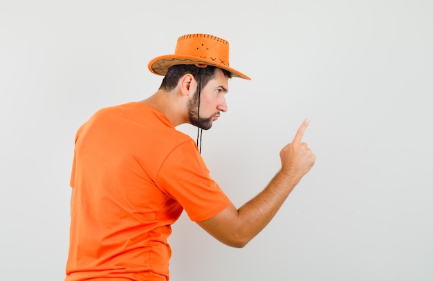 Foto gratuita hombre joven con camiseta naranja, sombrero advirtiendo a alguien con el dedo y mirando furioso.