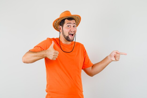 Hombre joven con camiseta naranja, sombrero apuntando hacia un lado, mostrando el pulgar hacia arriba y mirando feliz, vista frontal.