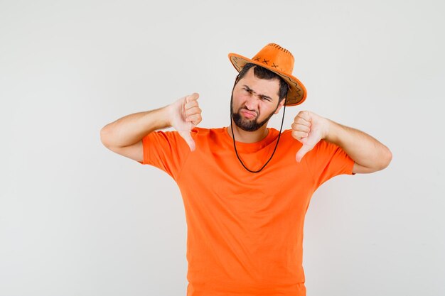 Hombre joven en camiseta naranja, sombrero mostrando el doble pulgar hacia abajo y mirando descontento, vista frontal.