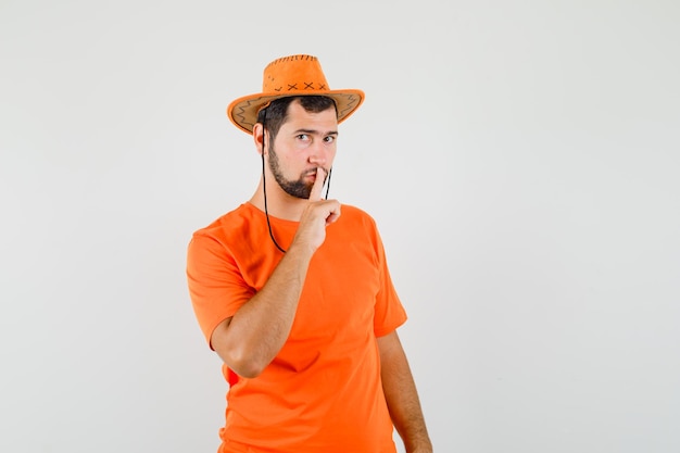 Hombre joven en camiseta naranja, sombrero mostrando gesto de silencio y mirando con cuidado, vista frontal.